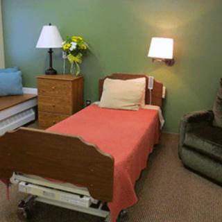 A hospital room with a single bed, side table, lamps, and a green chair.