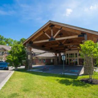 A building entrance with a large wooden canopy, surrounded by trees and a parking lot on a sunny day.