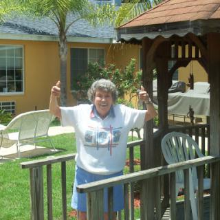 An elderly woman joyfully posing with her hands up in a sunny garden.