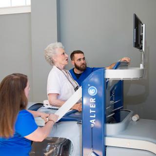 An elderly woman engaged in a rehabilitation session, assisted by two healthcare professionals using advanced technology.