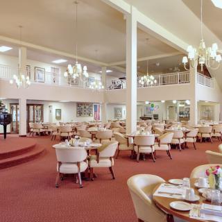 A spacious dining hall with elegantly arranged tables and chairs, featuring chandeliers and a carpeted floor.