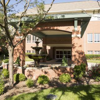 A sunny view of a building entrance with a fountain and landscaped garden.