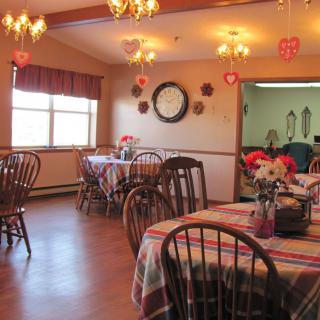 A cozy dining room with decorated tables and wooden chairs, featuring a large clock on the wall.