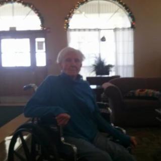 An elderly person sitting in a wheelchair indoors with large windows and furniture in the background.