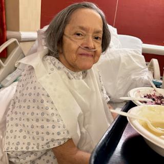 An elderly woman in a hospital bed enjoying her meal.