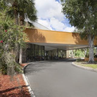 A modern building entrance surrounded by lush greenery and palm trees under a bright sky.