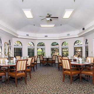 A bright dining room with multiple tables and chairs, featuring large windows and a ceiling fan.