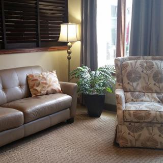 A cozy living room featuring a leather sofa, patterned armchair, lamp, and a potted plant by the window.