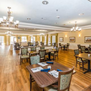 A spacious and well-lit dining room with neatly arranged tables and chairs, featuring elegant chandeliers and hardwood flooring.