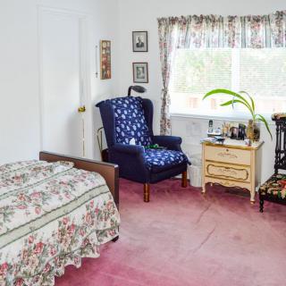 A cozy bedroom with a floral-patterned bedspread, blue armchair, and a wooden nightstand by the window.