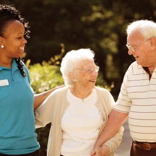 A caregiver assisting two elderly individuals outdoors, sharing a moment of happiness.