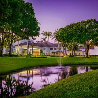 A tranquil evening view of a modern house with its reflection shimmering in a nearby pond, surrounded by lush green grass and trees.