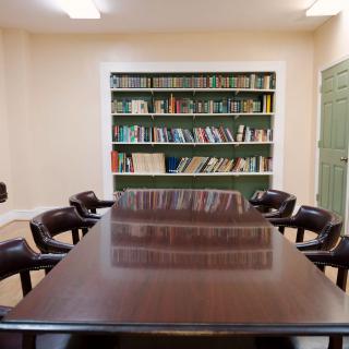 A conference room featuring a large wooden table surrounded by chairs, with a bookshelf in the background.
