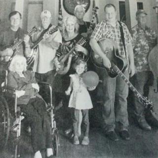 A black and white family photo featuring several people with musical instruments, including a guitar and banjo. An elderly person is seated in a wheelchair, and a child stands in the center holding a balloon.