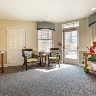 A cozy living room with sunlight streaming through large windows, featuring chairs and a bookshelf.