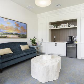 A modern living room with a stylish sofa, unique coffee table, and artistic wall painting. The space includes built-in shelving displaying decorative items, alongside a mini fridge and coffee maker.