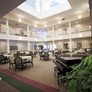 A spacious indoor atrium featuring multiple tables, chairs, a piano, and natural lighting from a skylight.