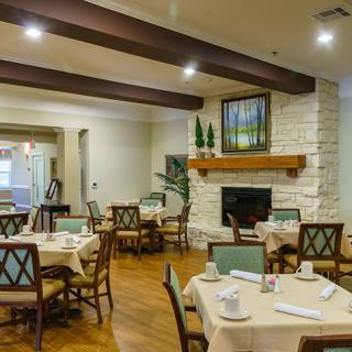 A cozy dining room with neatly set tables and a fireplace, complemented by wooden beams and soft lighting.