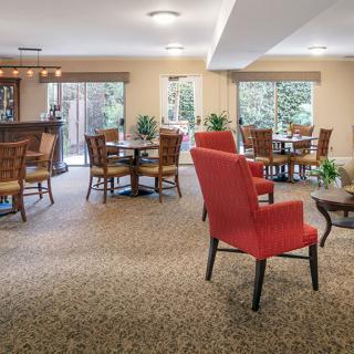 A cozy dining area with tables and red chairs arranged on a carpeted floor, surrounded by large windows and decorative plants.