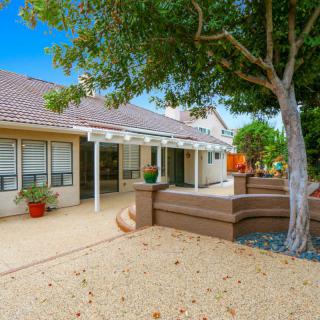 A serene backyard with a tree providing shade over a patio area, featuring a well-maintained garden.