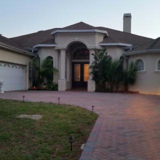 A view of a large, single-story house with a paved driveway and surrounding greenery.