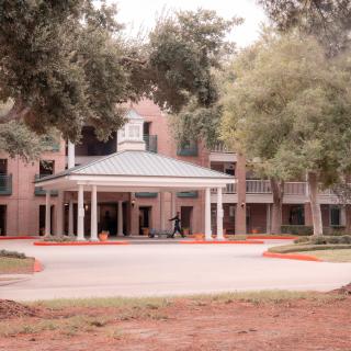 A two-story building with a porch and columns at the entrance, surrounded by trees and a driveway.