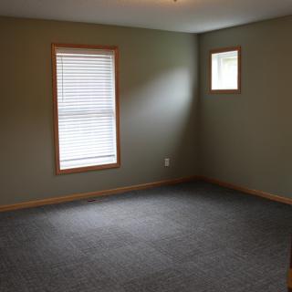 An empty room with carpet flooring and two windows allowing natural light.
