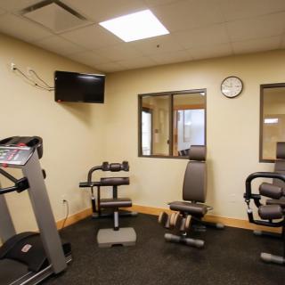 A small gym room with exercise equipment including a treadmill and strength training machines, featuring a wall-mounted television and clock.