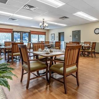 A well-lit dining room with wooden tables and chairs, featuring large windows and a chandelier.