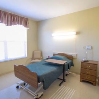 A hospital room with a single bed, bedside table, and a chair next to a large window.