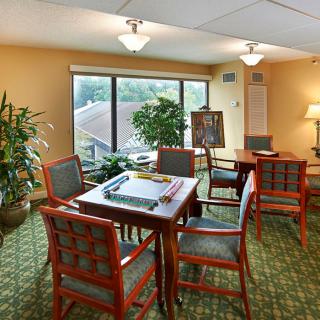 A comfortable, well-lit meeting area with a large window, featuring tables and chairs set for gathering or games, and decorated with plants.