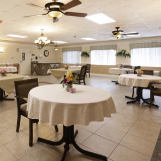 An empty dining room with round tables and chairs, featuring ceiling fans and natural light from large windows.