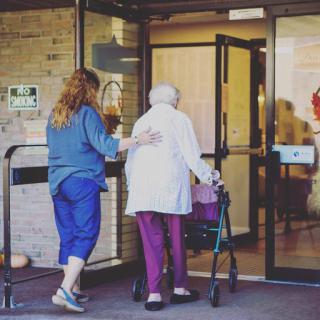 A caregiver assists an elderly woman using a walker as they enter a building.
