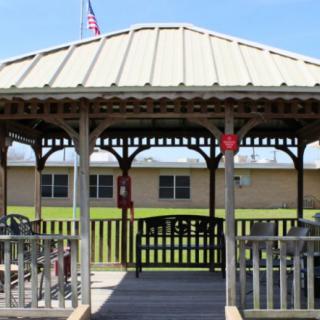 A wooden gazebo with a metal roof in front of a building and an American flag in the background.