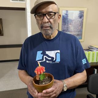 A man wearing a hat and glasses holds a potted plant indoors.