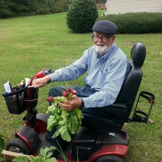 An elderly man in a cap gathers vegetables while sitting on a mobility scooter in his garden.