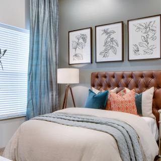 A cozy bedroom with a tufted headboard, framed botanical artwork, and layered bedding featuring various textured pillows.