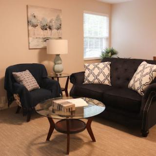 A cozy living room setup featuring a black couch with patterned cushions, a comfortable blue armchair, and a stylish glass coffee table.