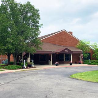 A charming brick building surrounded by lush greenery and trees, with a winding driveway leading to its entrance.