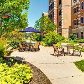 A tranquil outdoor patio area with chairs and a table under an umbrella, surrounded by lush greenery and adjacent to a large building.
