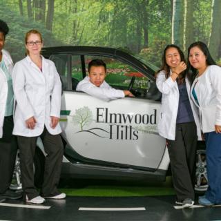 A group of people in white lab coats standing around a small car with 'Elmwood Hills' branding, indoors.