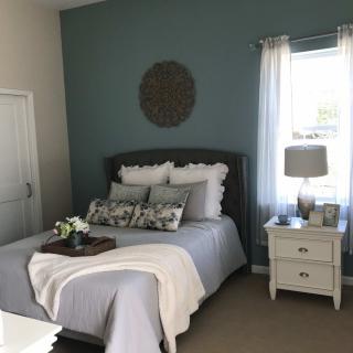 A cozy bedroom with a neatly made bed, pillows, and a soft blanket. A nightstand with a lamp sits beside the bed under a sunlit window.