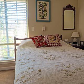 A cozy bedroom featuring a quilt-covered bed with decorative pillows, next to a window with blinds, and furnished with a lamp and mirror.