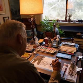 An artist working on a painting in a cozy studio filled with art supplies, books, and plants by the window.