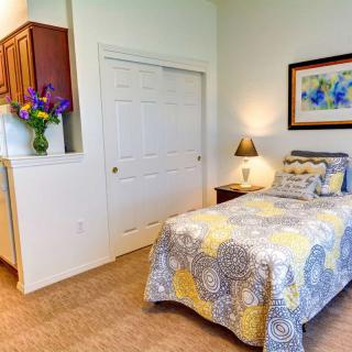 A cozy bedroom with a single bed featuring a colorful duvet, a bedside lamp, and decorative elements like flowers and a painting.