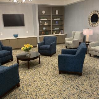 A cozy living room setup with blue and gray chairs, a central table with flowers, a TV on the wall, and decorative shelves.