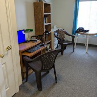 A small office space featuring a desk with a computer, two plastic chairs, and a bookshelf near a window.