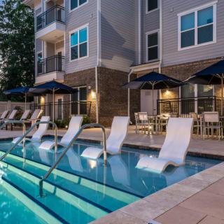 A modern apartment complex features a relaxing swimming pool area with lounge chairs and umbrellas, set against an evening backdrop.