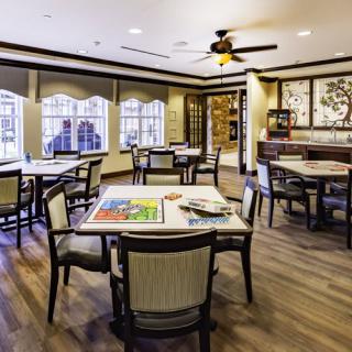 A cozy dining room interior with multiple tables and chairs, featuring large windows and a ceiling fan.