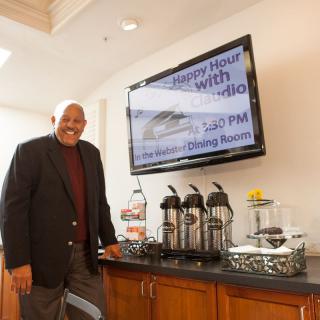 A man in business attire stands smiling near a coffee station with a sign advertising happy hour.